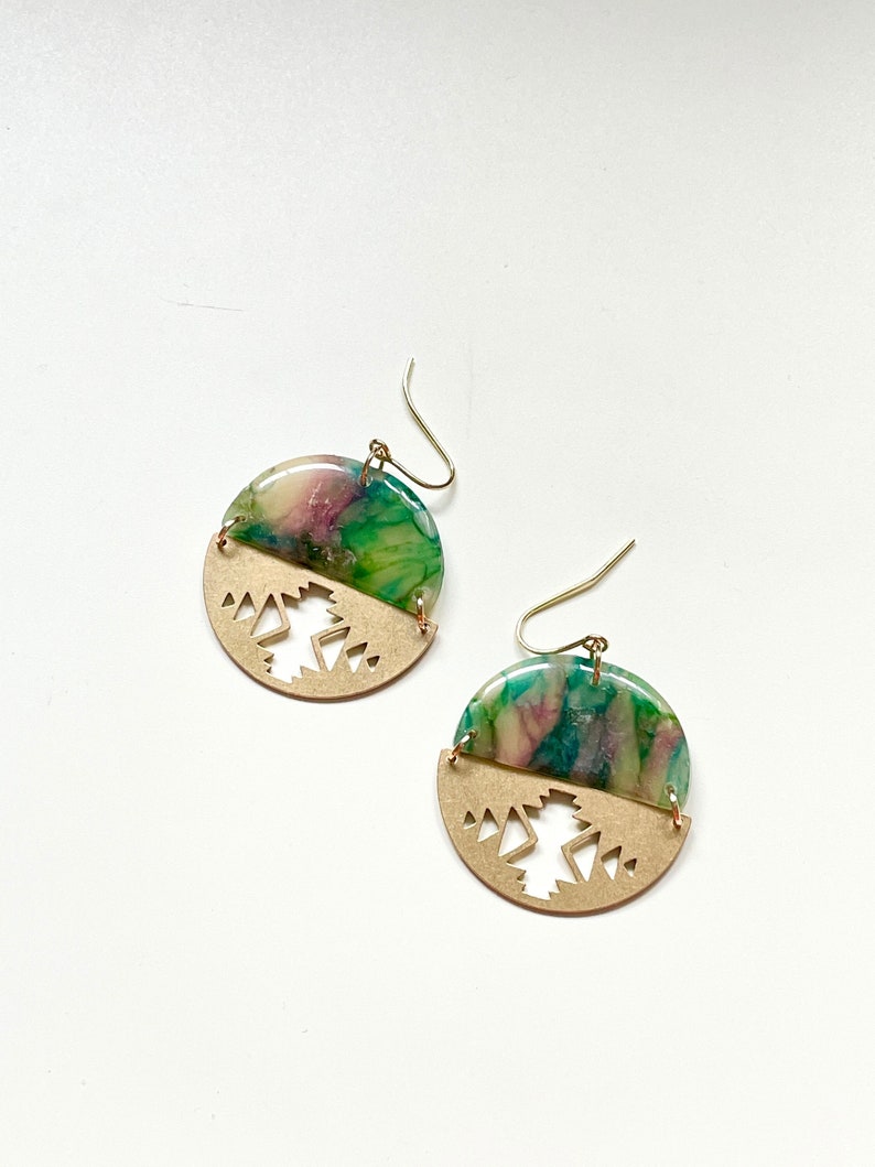 May include: A pair of handcrafted earrings. The top half features a marbled design in shades of green, pink, and purple. The bottom half is a gold-colored metal with a geometric cutout design. Gold-colored ear wires complete the earrings.