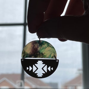 May include: A unique earring with a half-circle design. The top half features a marbled pattern with green and purple hues, while the bottom half is a dark metal with a geometric cutout design. The earring is held up by a small hook.