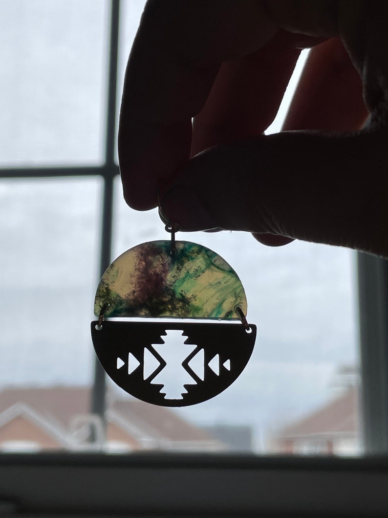 May include: A unique earring with a half-circle design. The top half features a marbled pattern with green and purple hues, while the bottom half is a dark metal with a geometric cutout design. The earring is held up by a small hook.