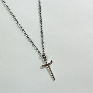 May include: A silver necklace featuring a small sword pendant. The pendant is a detailed miniature of a sword, with a pointed blade and a cross-shaped hilt. The chain is a delicate link style, and the necklace is displayed against a white background.