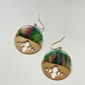 May include: A pair of handmade earrings. Each earring features a half-circle of gold-colored metal with a geometric cutout design, and a half-circle of marbled green, pink, and cream-colored resin. Gold-colored ear wires complete the design.