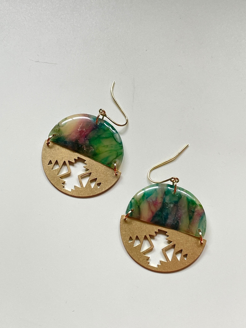 May include: A pair of handmade earrings. Each earring features a half-circle of gold-colored metal with a geometric cutout design, and a half-circle of marbled green, pink, and cream-colored resin. Gold-colored ear wires complete the design.