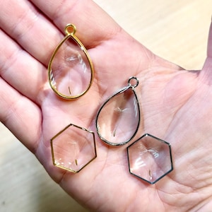 May include: Four clear glass pendants, each containing a dandelion seed. Two are teardrop-shaped, one with a gold frame and one with a silver frame. The other two are hexagonal, one with a gold frame and one with a silver frame.