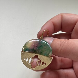 May include: Round earrings with a half-circle design. The top half is a mix of green, purple, and yellow hues, while the bottom half is a gold-colored metal with a geometric mountain design. The earrings have gold-colored hooks.