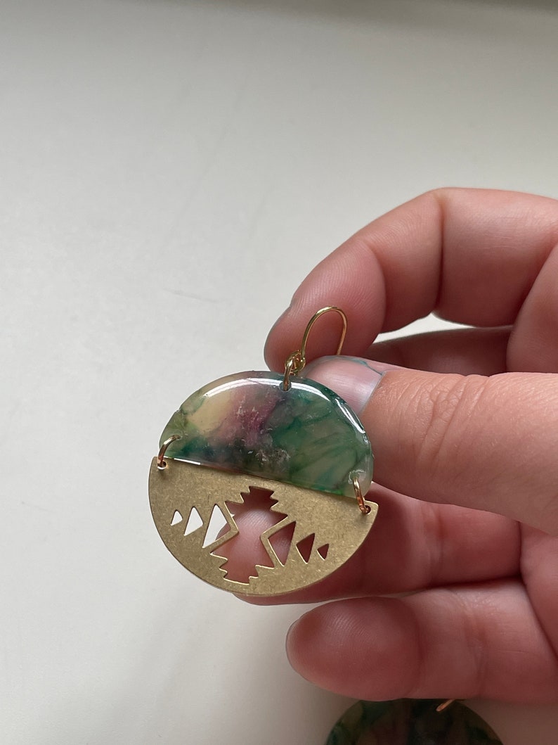 May include: Round earrings with a half-circle design. The top half is a mix of green, purple, and yellow hues, while the bottom half is a gold-colored metal with a geometric mountain design. The earrings have gold-colored hooks.