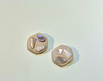 May include: Two beige, hexagonal stud earrings. Each earring is inlaid with iridescent, shell-like fragments. The earrings are set against a plain, light background. These earrings are a unique accessory.