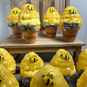May include: A set of six yellow and black honey pot themed decorations with a bee on top. Each pot has a black and white checkered ribbon and the word "Hunny" written on the front.
