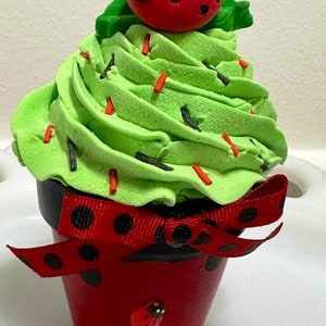 May include: A green cupcake with red and black sprinkles topped with a red ladybug. The cupcake is in a red pot with black polka dots and a red ribbon.