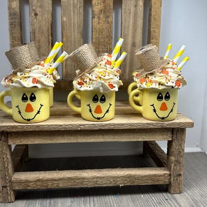 May include: Three yellow mugs with a scarecrow face design, topped with whipped cream, sprinkles, and a straw. Each mug has a small burlap hat. The mugs are arranged on a rustic wooden bench.