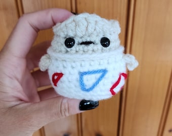 May include: A handmade crochet Togepi plushie. The plushie features a cream-colored head and a white body with red and blue accents. It has black button eyes and small arms. The Togepi is a cute, whimsical character.