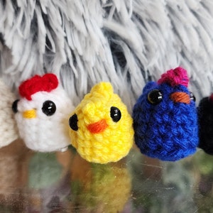 May include: Five crocheted chicks in different colors, including white, yellow, blue, and black. Each chick has a red comb and black eyes.