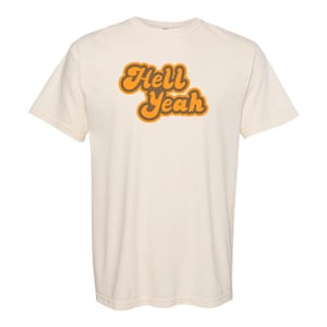 May include: A cream colored t-shirt with the text "Hell Yeah" in orange and brown retro lettering.