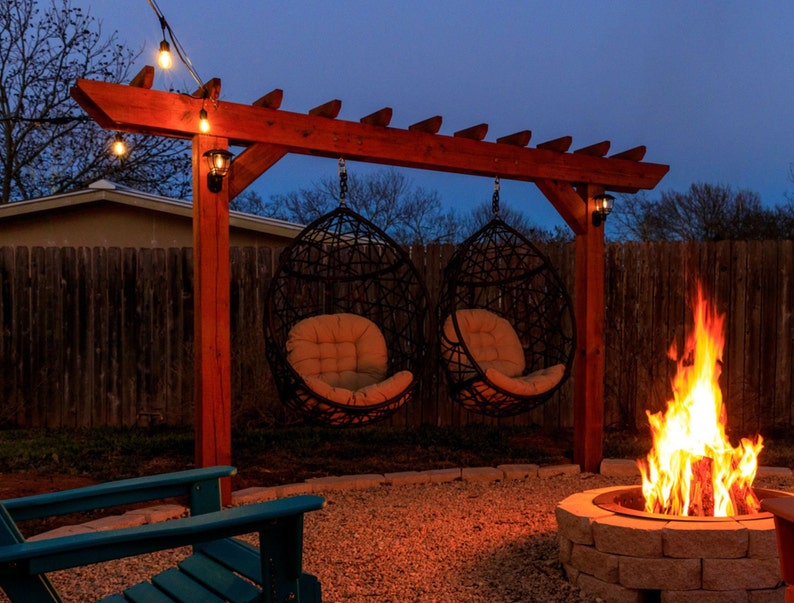 May include: Outdoor scene at dusk featuring a wooden pergola with two hanging egg chairs. A fire pit with a burning fire is in the foreground. String lights are strung across the pergola, and the scene is set against a wooden fence.