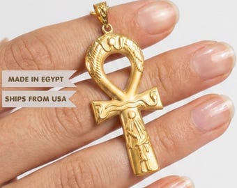 May include: A gold-colored metal ankh pendant featuring an embossed figure and snake motifs. The pendant has a loop at the top for a chain. Text on the image reads: MADE IN EGYPT and SHIPS FROM USA. The design is symmetrical with a cross-like shape.