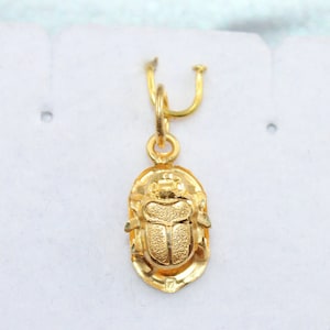 May include: A gold pendant shaped like a scarab beetle with a textured, matte finish on the wings and body. The pendant features a small circular jump ring at the top for attachment, showcasing a detailed insect design in a polished gold tone.