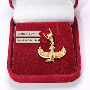 May include: A gold-coloured pendant featuring a winged figure, likely representing an Egyptian deity, with outstretched wings and a kneeling posture. The pendant is displayed inside an open, rectangular, red velvet-lined jewellery box.
