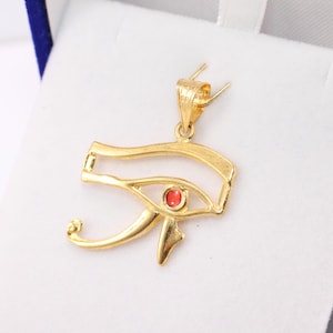 May include: Gold-coloured Eye of Horus pendant, featuring a small red gemstone. The pendant is presented in a blue velvet jewellery box with a white interior. The pendant is a symbol of protection and good health, often worn as an amulet.