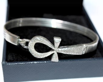 May include: A silver-colored bracelet with an Ankh symbol in the center, resting inside a black box. The Ankh is a cross-like symbol with a loop at the top, often associated with ancient Egyptian culture. The bracelet is a bangle style.