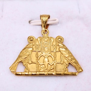 Può includere: Un ciondolo color oro raffigurante due uccelli simmetrici rivolti verso un cartiglio centrale. Il design include simboli ankh e una figura seduta alla base. Il ciondolo presenta una contromaglia testurizzata per l'inserimento di una catena.