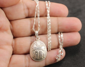 May include: A silver-colored necklace featuring an oval-shaped pendant with an embossed scarab beetle design. The pendant hangs from a delicate silver-toned chain with a small metal clasp visible at the end, showcasing a metallic finish.