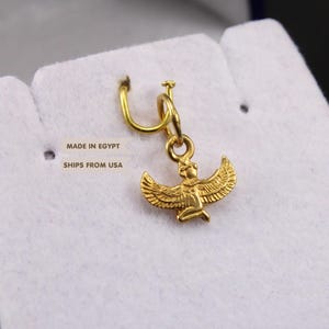 May include: A gold-colored metal charm featuring a winged figure with outstretched wings, suspended from a small circular jump ring. The charm displays intricate detailing on the wings and body, presented against a white textured surface.