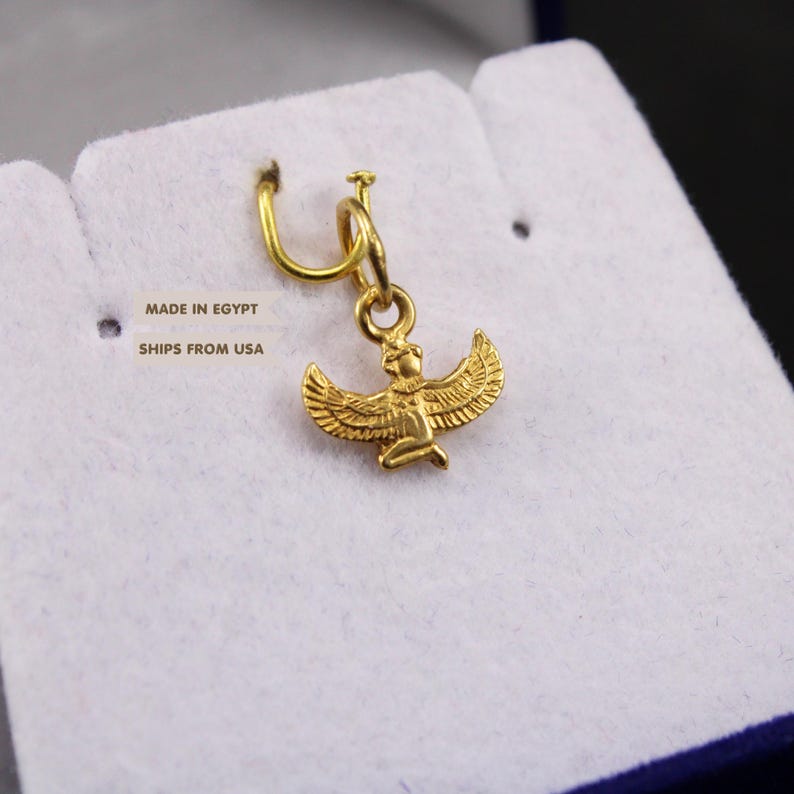 May include: A gold-colored metal charm featuring a winged figure with outstretched wings, suspended from a small circular jump ring. The charm displays intricate detailing on the wings and body, presented against a white textured surface.