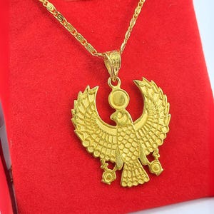 Può includere: Un ciondolo color oro raffigurante un falco stilizzato con le ali spiegate, sormontato da un disco solare circolare. Il ciondolo è sospeso a una catena a maglie color oro abbinata, che mette in risalto dettagli di piume incise.