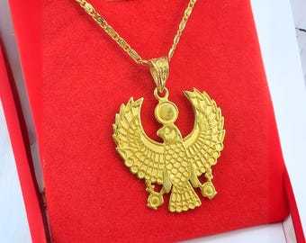 May include: A gold-colored pendant necklace featuring an Egyptian Horus design. The pendant, shaped like a bird with outstretched wings, is displayed on a red background. The necklace chain is also gold-colored.