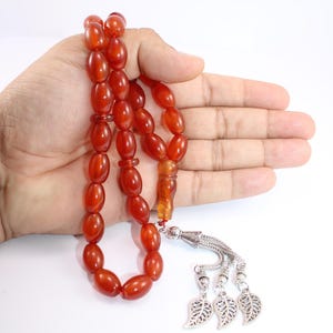 May include: A strand of reddish-orange prayer beads with oval-shaped beads. The beads are held in a hand against a white background. The end of the strand features a decorative silver-colored tassel with leaf-shaped charms.