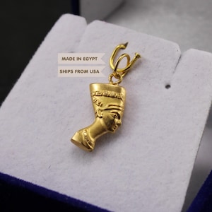 May include: A gold-colored pendant featuring the profile of Queen Nefertiti wearing a tall crown. The pendant is attached to a small gold-colored jump ring. Text visible on the image includes "MADE IN EGYPT" and "SHIPS FROM USA".