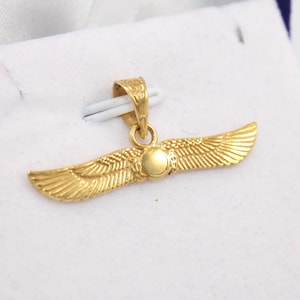 May include: A gold-coloured pendant with a wing design. The pendant features outstretched wings with detailed feathering and a central circular element. The pendant is displayed on a white surface.
