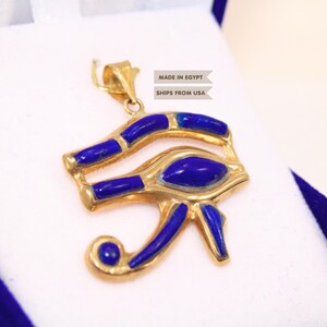 May include: A gold-toned Eye of Horus pendant featuring inlaid deep blue stone segments. The pendant includes a metal bail at the top for attachment. The text MADE IN EGYPT and SHIPS FROM USA is visible near the top of the pendant design.