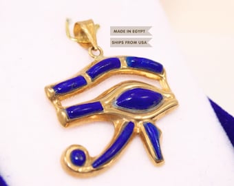 Puede incluir: Un colgante dorado con forma del Ojo de Horus que presenta incrustaciones de lapislázuli azul intenso. El colgante tiene una anilla de metal en la parte superior. El diseño es una representación estilizada del antiguo símbolo egipcio con piedras azules.