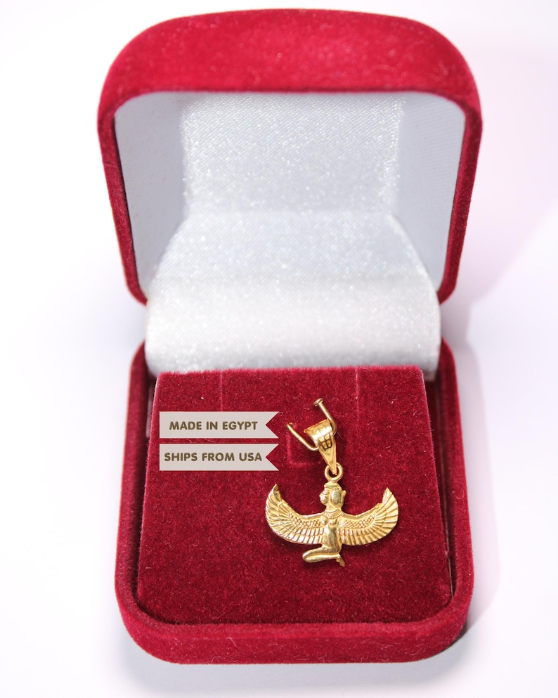 May include: A gold-coloured pendant featuring a winged figure, likely representing an Egyptian deity, with outstretched wings and a kneeling posture. The pendant is displayed inside an open, rectangular, red velvet-lined jewellery box.