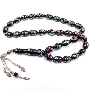Peut inclure: Un chapelet de prière noir avec des perles rouges et turquoise. Les perles ovales sont décorées de points blancs. Le collier a un pompon argenté et une poignée décorative.