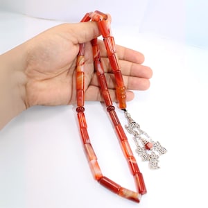 May include: Red and orange prayer beads with a silver tassel. The rectangular beads have a marbled appearance. The ornate tassel features small charms. The beads are held in a hand against a white background.