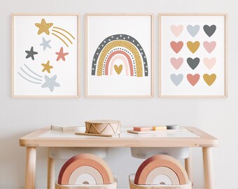 Könnte beinhalten: Drei gerahmte Drucke mit einem Regenbogen, Sternen und Herzen. Der Regenbogen ist in den Farben Rosa, Orange, Gelb und Blau gehalten. Die Sterne sind gelb und blau. Die Herzen sind rosa, rot und gold. Die Drucke hängen an einer weißen Wand über einem Holztisch mit zwei regenbogenförmigen Hockern.