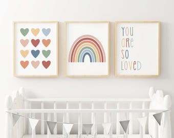 Könnte beinhalten: Drei gerahmte Drucke mit einem Regenbogen, Herzen und dem Text "You are so loved". Die Drucke hängen über einem weißen Kinderbett mit einer grauen Dreiecksgirlande.