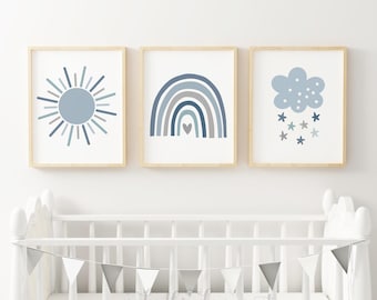 Könnte beinhalten: Drei gerahmte Drucke mit einer blauen Sonne, einem Regenbogen mit einem Herzen und einer blauen Wolke mit Sternen. Die Drucke hängen an einer weißen Wand über einem weißen Kinderbett mit einer grau-weißen Girlande.