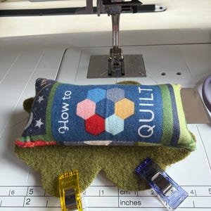 May include: A rectangular fabric pincushion with the text How to QUILT and a colorful hexagonal pattern, resting on a green felt base. Two plastic sewing clips in yellow and purple are placed on the surface next to a ruler showing inches and cm.