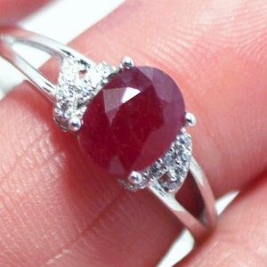 May include: A silver ring featuring a large, red ruby gemstone surrounded by smaller, clear gemstones.