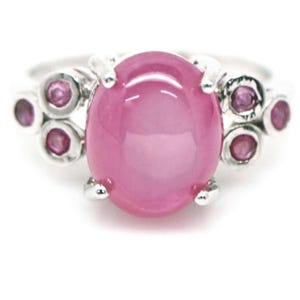 May include: A silver ring with a large, oval pink gemstone at its centre. Smaller, round pink gemstones are set on either side of the main stone. The ring has a bright, polished finish.