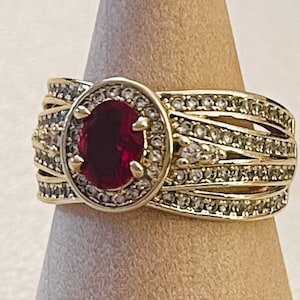 May include: A gold ring featuring a central oval-cut deep red gemstone surrounded by a halo of small clear stones. The band is composed of multiple intersecting gold strands, each encrusted with rows of small clear stones, creating a wide design.