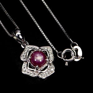 Könnte beinhalten: Eine silberne Halskette mit einem blütenförmigen Anhänger. Der Anhänger hat einen zentralen, runden, purpurroten Edelstein, umgeben von kleinen, klaren Steinen. Kette und Verschluss sind silberfarben, vor schwarzem Hintergrund.