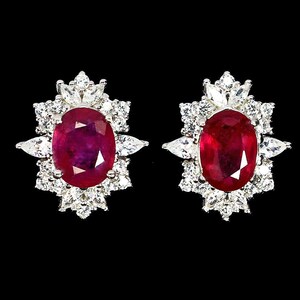 May include: A pair of stud earrings featuring oval-cut, deep red rubies surrounded by a halo of small, sparkling white gemstones. The earrings have a floral-like design, set against a black background.