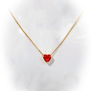 May include: A gold-colored necklace with a small, heart-shaped, red gemstone pendant. The pendant is centered on a delicate chain, creating a simple and elegant design. The necklace is displayed against a white background.