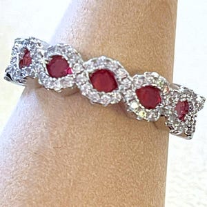 May include: A silver ring featuring a series of vibrant red gemstones, each encircled by a halo of smaller, sparkling white stones. The ring is displayed on a neutral-toned surface, showcasing its elegant design.