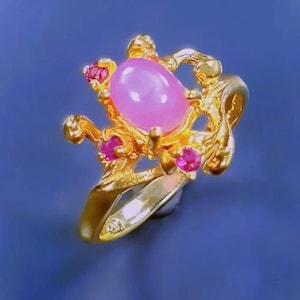 Könnte beinhalten: Ein goldener Ring mit einem ovalen rosafarbenen Edelstein, der von kleineren rosafarbenen Edelsteinen umgeben ist. Der Ring hat ein zartes, verziertes Design.