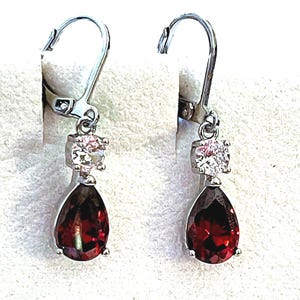 May include: A pair of silver-toned drop earrings featuring a clear, round-cut stone at the top and a larger, pear-shaped, deep red stone at the bottom. The earrings are secured with a lever-back closure mechanism for a secure fit.