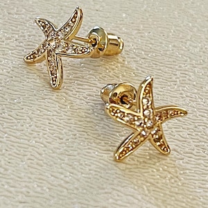 May include: A pair of gold-toned stud earrings shaped like starfish. Each earring features a textured surface embedded with small, clear, sparkling stones along the five arms. The earrings include matching gold-toned post backs for secure fastening.